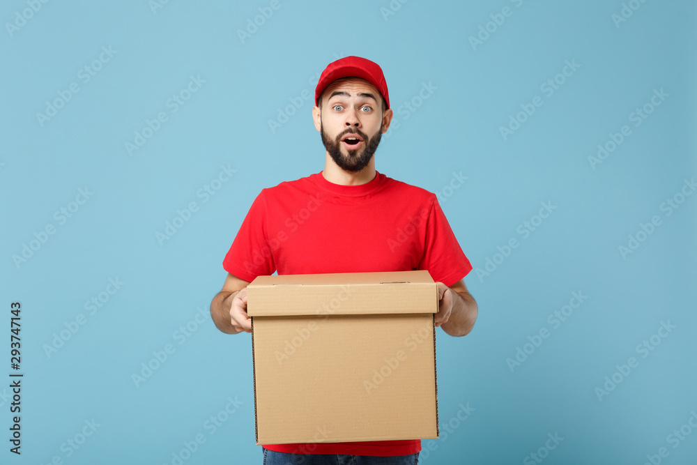 Delivery man in red uniform isolated on blue background, studio portrait. Male employee in cap t-shirt print working as courier dealer hold empty cardboard box. Service concept. Mock up copy space. Delivery man in red uniform isolated on blue background, studio portrait. Male employee in cap t-shirt print working as courier dealer hold empty cardboard box. Service concept. Mock up copy space.