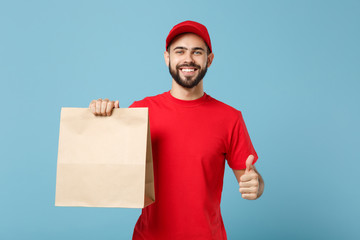 Delivery man in red uniform hold craft paper packet with food isolated on blue background, studio portrait. Male employee in cap t-shirt print working as courier. Service concept. Mock up copy space.