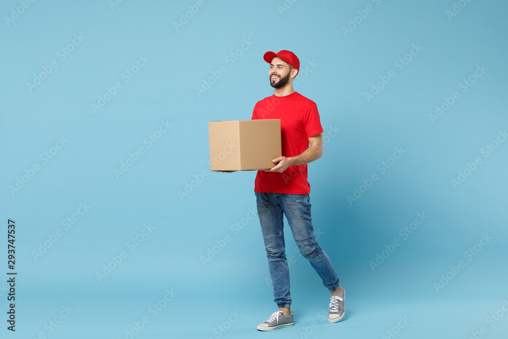 Delivery man in red uniform isolated on blue background, studio portrait. Male employee in cap t-shirt print working as courier dealer hold empty cardboard box. Service concept. Mock up copy space. Delivery man in red uniform isolated on blue background, studio portrait. Male employee in cap t-shirt print working as courier dealer hold empty cardboard box. Service concept. Mock up copy space.