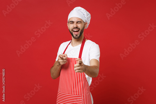 Bearded male chef cook or baker man in striped apron t-shirt toque chefs hat posing isolated on red background. Cooking food concept. Mock up copy space. Blinking, pointing index fingers on camera.