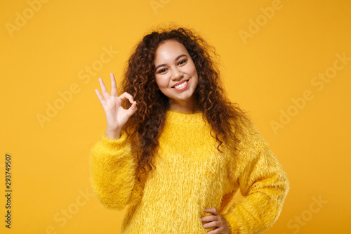 Smiling young african american girl in fur sweater posing isolated on yellow orange wall background, studio portrait. People sincere emotions lifestyle concept. Mock up copy space. Showing OK gesture. © ViDi Studio