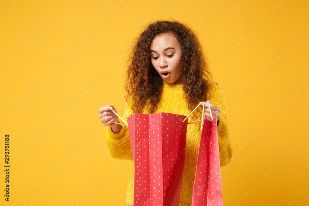 Shocked young african american girl in fur sweater posing isolated on yellow orange wall background. People lifestyle concept. Mock up copy space. Holding package bag with purchases after shopping. Shocked young african american girl in fur sweater posing isolated on yellow orange wall background. People lifestyle concept. Mock up copy space. Holding package bag with purchases after shopping.