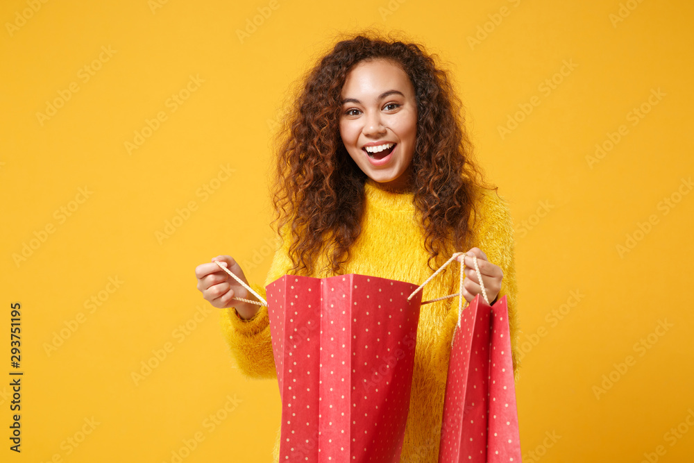 Surprised young african american girl in fur sweater posing isolated on yellow orange wall background. People lifestyle concept. Mock up copy space. Holding package bag with purchases after shopping. Surprised young african american girl in fur sweater posing isolated on yellow orange wall background. People lifestyle concept. Mock up copy space. Holding package bag with purchases after shopping.