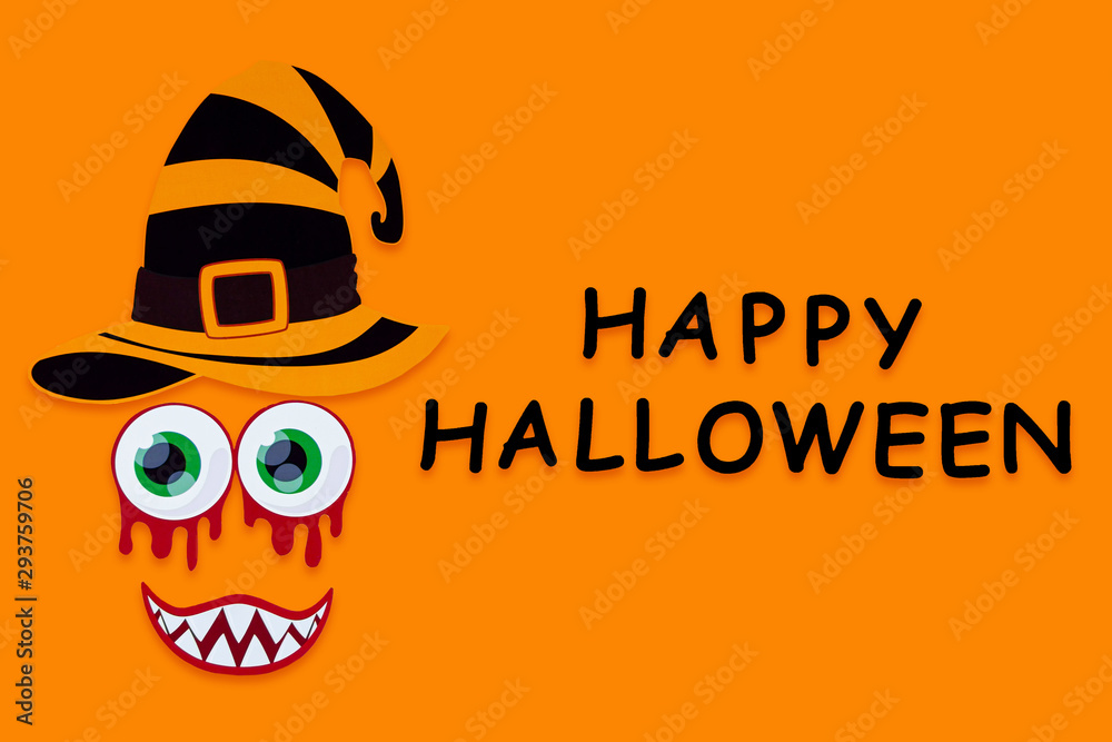 Obraz premium Funny face of witch monster with blood eyes and vampire smile on orange background. Paper decor, photo props with black inscription happy halloween on canvas. Party accessories for celebration.