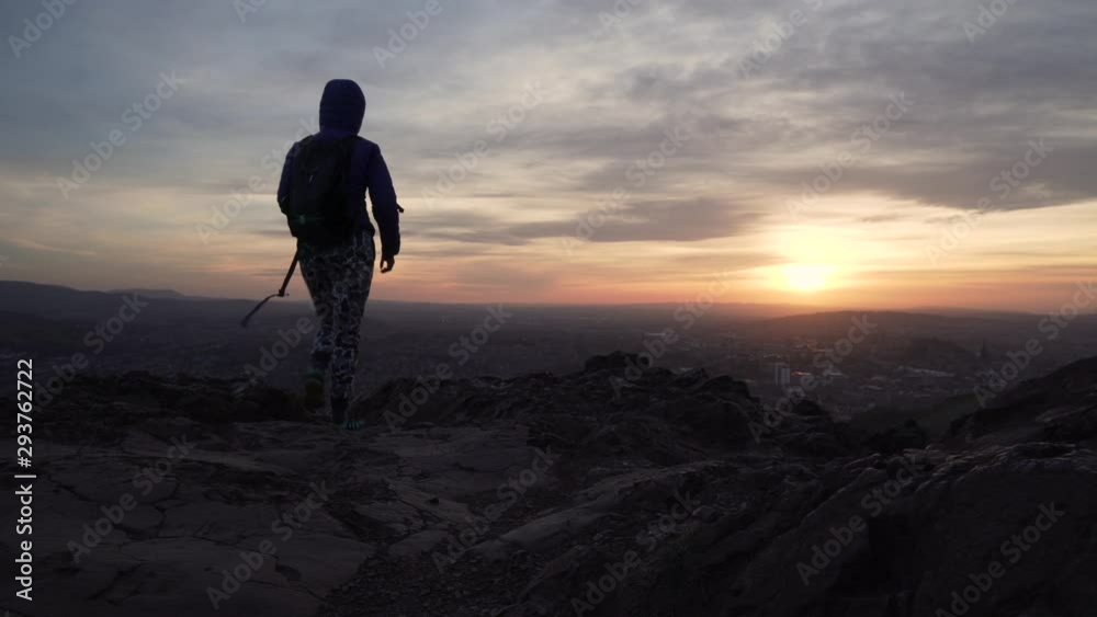 Static shot of girl walking to the edge of a mountain to take a photo with smartphone during beautiful sunset of Edinburgh cityscape with rocky cliffs in foreground Static shot of girl walking to the edge of a mountain to take a photo with smartphone during beautiful sunset of Edinburgh cityscape with rocky cliffs in foreground