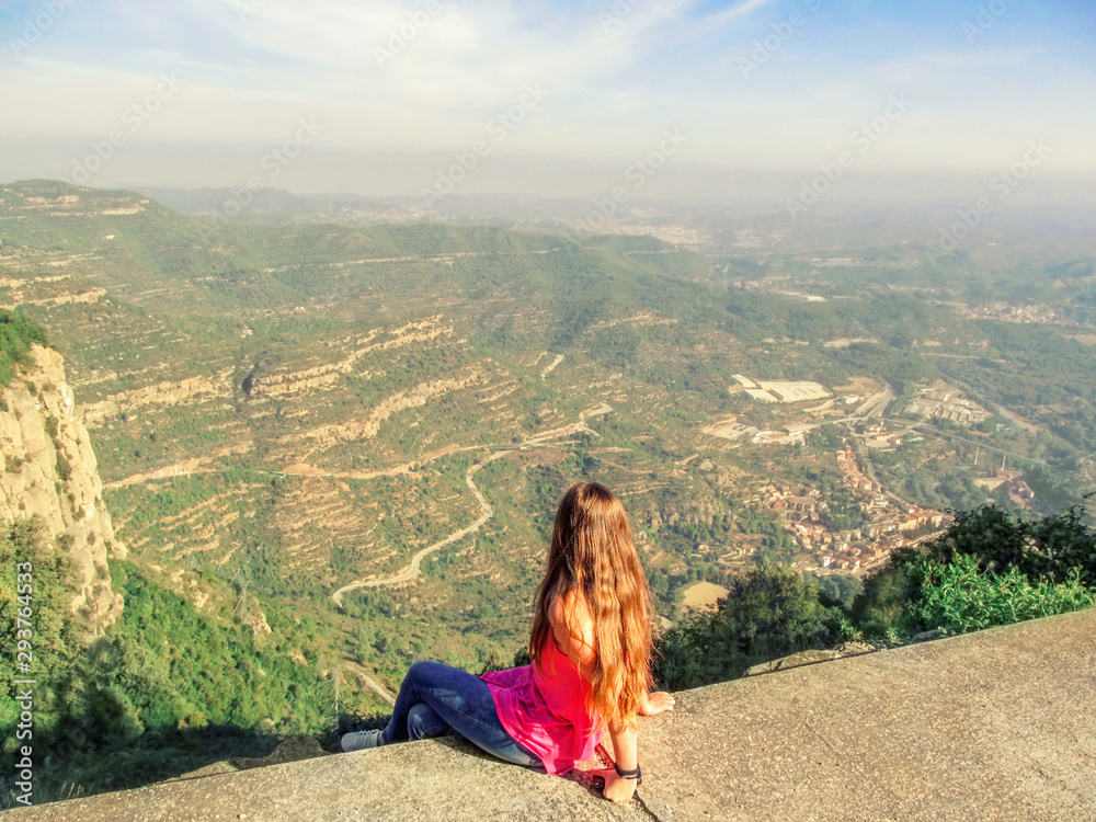 Blonde young woman sits on a viewing platform and looks down at Montserrat Mountain valley in Catalonia (Spain), view from the back. Beautiful landscape with fog on horizon, background with copy space