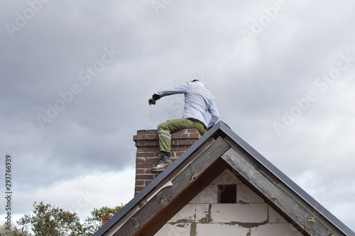 Konstfotografi Chimney sweep man cleaning brown brick chimney while sitting on chimney on building roof on cloudy sky background with copy space for text