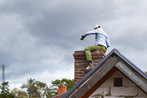 Tela Chimney sweep man cleaning brown brick chimney while sitting on chimney on building roof on cloudy sky background with copy space for text