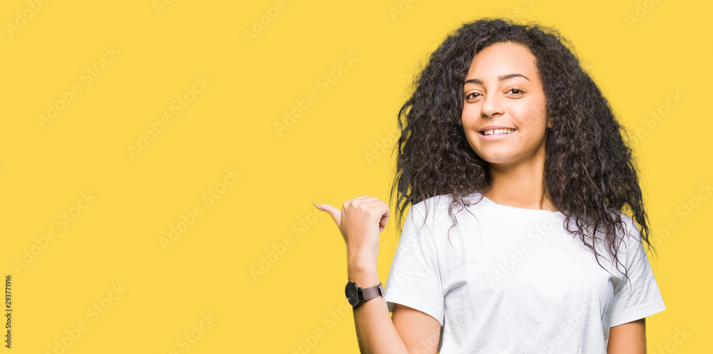 Young beautiful girl with curly hair wearing casual white t-shirt smiling with happy face looking and pointing to the side with thumb up. Young beautiful girl with curly hair wearing casual white t-shirt smiling with happy face looking and pointing to the side with thumb up.