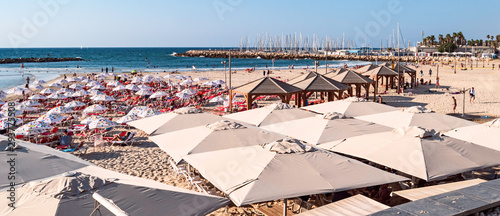 tourists enjoying the beach and sun shade umbrellas at gordon beach in Tel Aviv with the marina and sailboats in the background