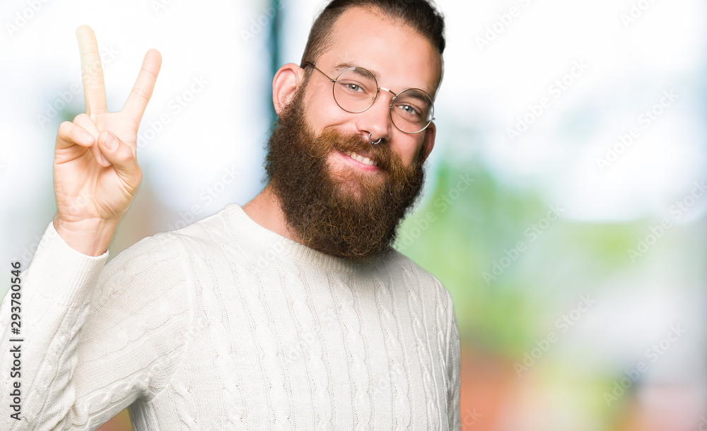 Young hipster man wearing glasses and winter sweater smiling with happy face winking at the camera doing victory sign. Number two. Young hipster man wearing glasses and winter sweater smiling with happy face winking at the camera doing victory sign. Number two.
