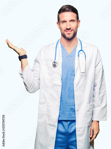 Handsome doctor man wearing medical uniform over isolated background smiling cheerful presenting and pointing with palm of hand looking at the camera.