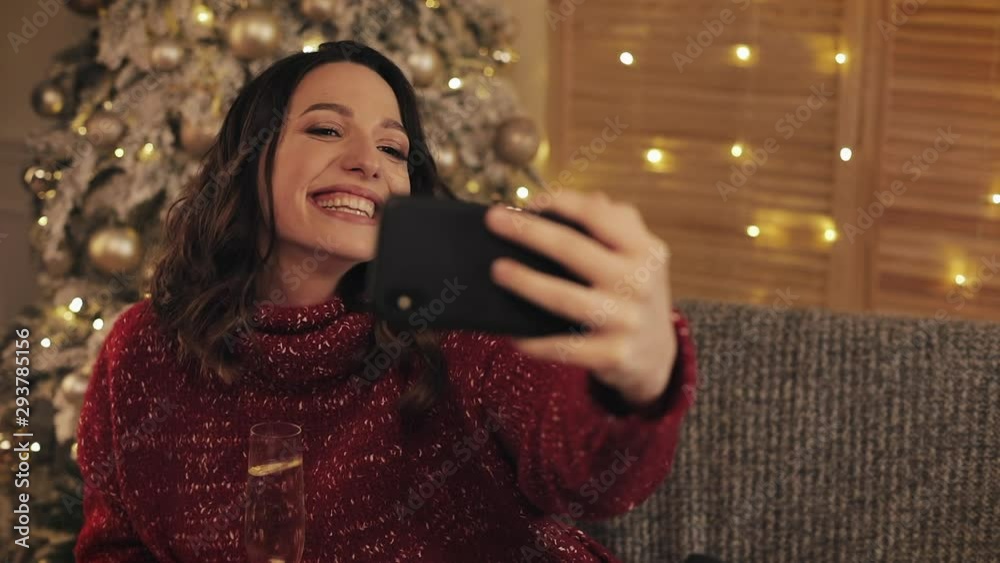 Beautiful Young Smiling Brunette Woman Raising Champagne Glass Sitting near Christmas Tree with Shining Lights at the Background Making Video Message or Selfie Concept of Holidays and New Year.