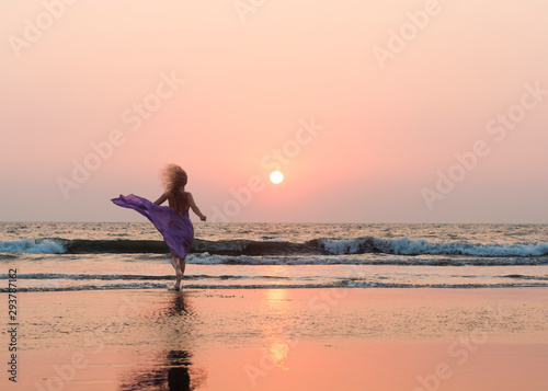 Beautiful young woman in a long purple dress running to sunset sea. Beach holidays travel concept