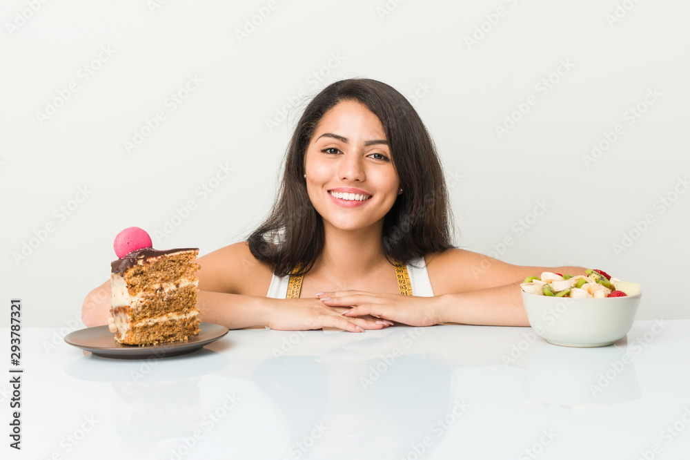 Young hispanic woman choosing between cake or fruit happy, smiling and cheerful. Young hispanic woman choosing between cake or fruit happy, smiling and cheerful.