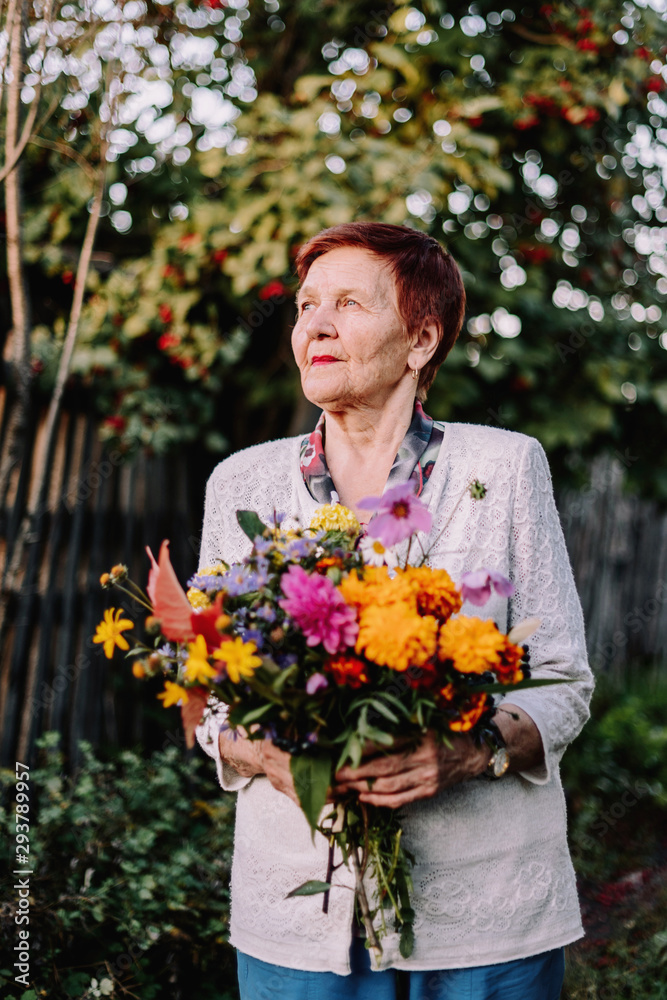 Fototapeta premium Grandmother cute smiles with flowers in her hands. Elderly woman portrait. Bouquet of flowers in the fall.