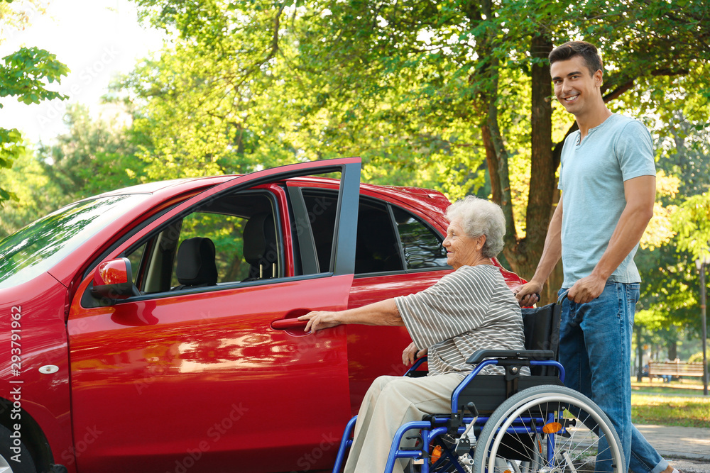 Young man helping disabled senior woman in wheelchair to get into car outdoors