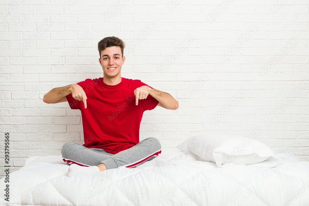 Fototapeta premium Young teenager student man on the bed points down with fingers, positive feeling.