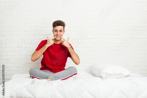 Young teenager student man on the bed smiles, pointing fingers at mouth.