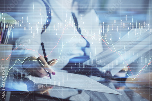 A woman hands writing information about stock market in notepad. Forex chart holograms in front. Concept of research.