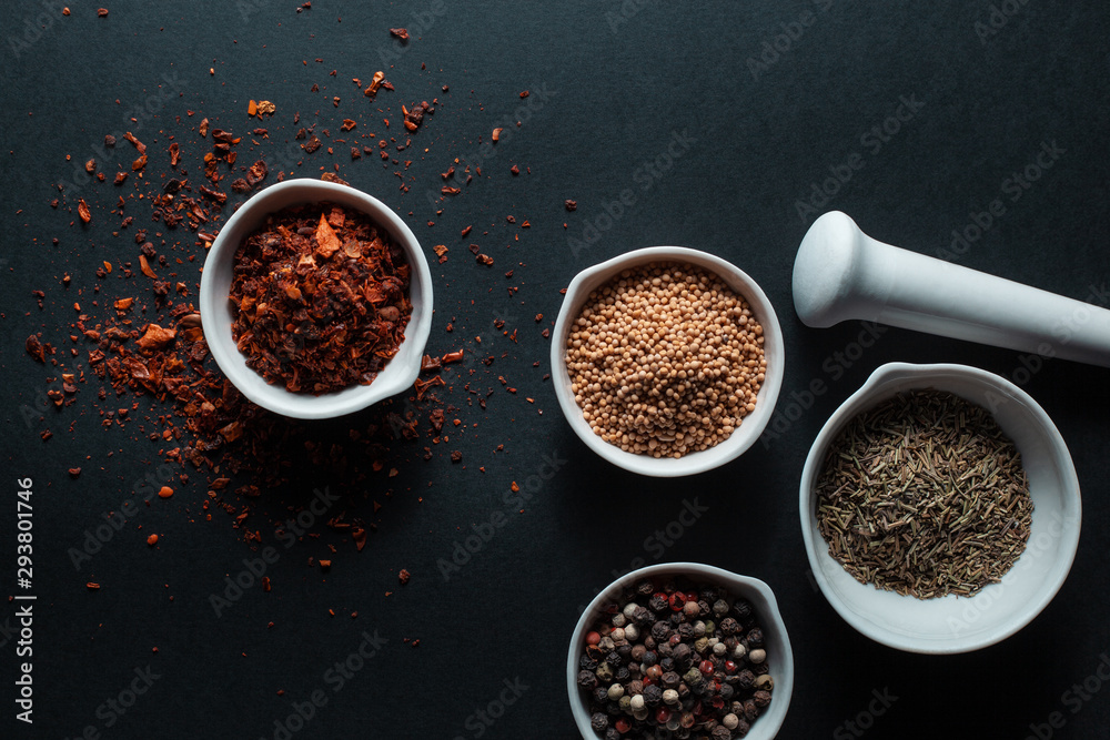 Paprika, mustard, caraway seeds and a mixture of peppercorns in white ceramic bowls next to the mortar isolated on black background