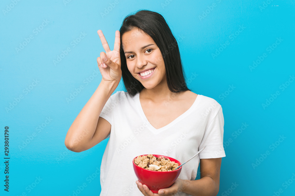 Young hispanic woman holding a cereal bowl showing victory sign and smiling broadly. Young hispanic woman holding a cereal bowl showing victory sign and smiling broadly.