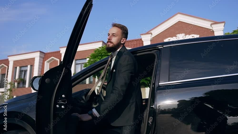 Handsome young caucasian businessman in formal suit with tie gets out of the black car and accepts an important phone call.