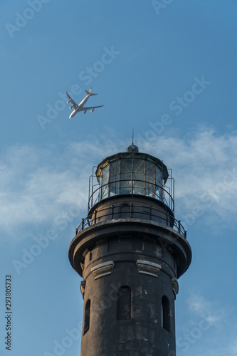 Flying passenger jet airplane passes over lighthouse monument on clear summer day past blue sky and clouds. Light houses used for aviation and maritime navigation with circular strobe spot light