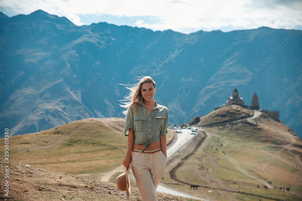 Naklejka premium Outdoor travel lifestyle smiling woman tourist posing against the backdrop of the mountains and medieval church / monastery. Safari style girl with hat