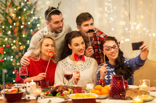 holidays and celebration concept - happy friends with party props taking selfie by smartphone at home christmas dinner