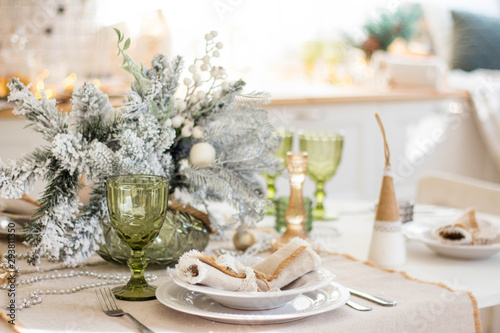 Beautiful served table with decorations, candles with glaze on white tablecloth on the background of the kitchen with people. Living room decorated with lights and Christmas tree.