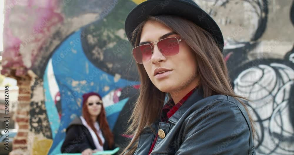 Close up of the young beautiful charming girl in hipster style, sunglasses and black hat looking at the camera and posing to it like a model at the graffity on the wall in slums. Outside. Portrait.