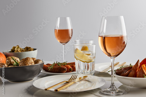 glasses with rose wine and lemon water, baked potatoes and carrots, white plates and cutlery on marble table isolated on grey