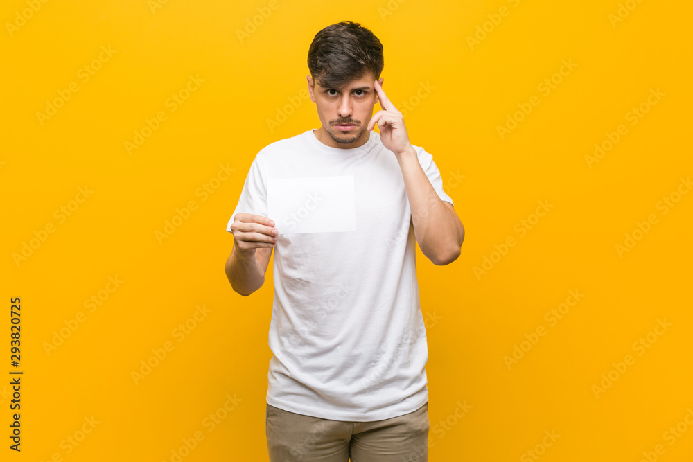 Young hispanic man holding a placard pointing his temple with finger, thinking, focused on a task. Young hispanic man holding a placard pointing his temple with finger, thinking, focused on a task.