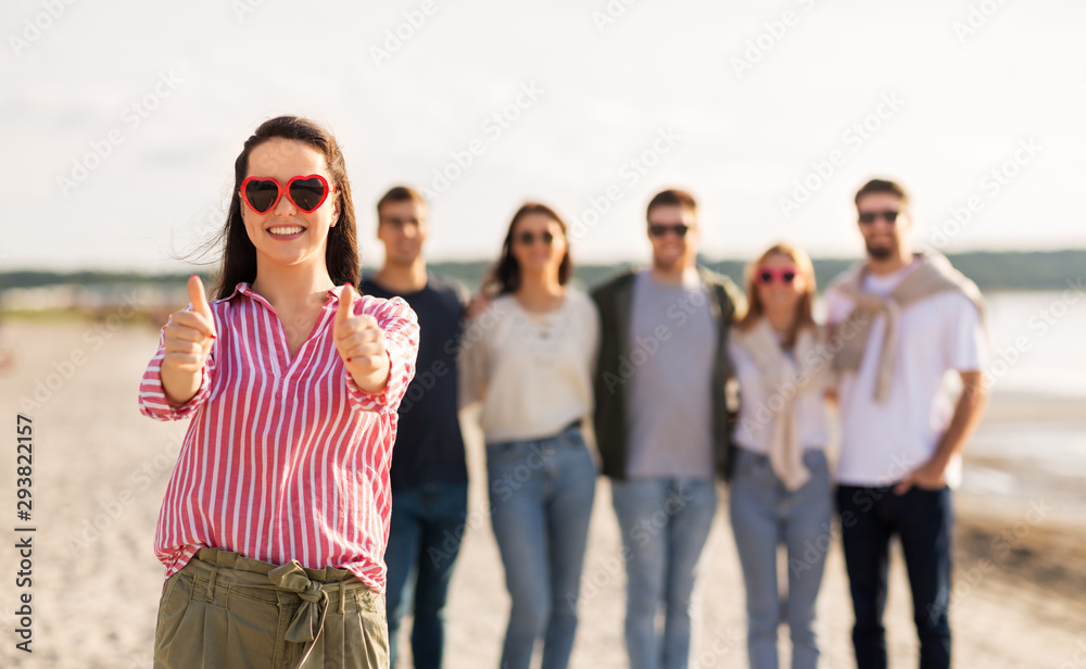 friendship, valentine's day and people concept - happy woman in heart-shaped sunglasses with group of friends on beach in summer showing thumbs up