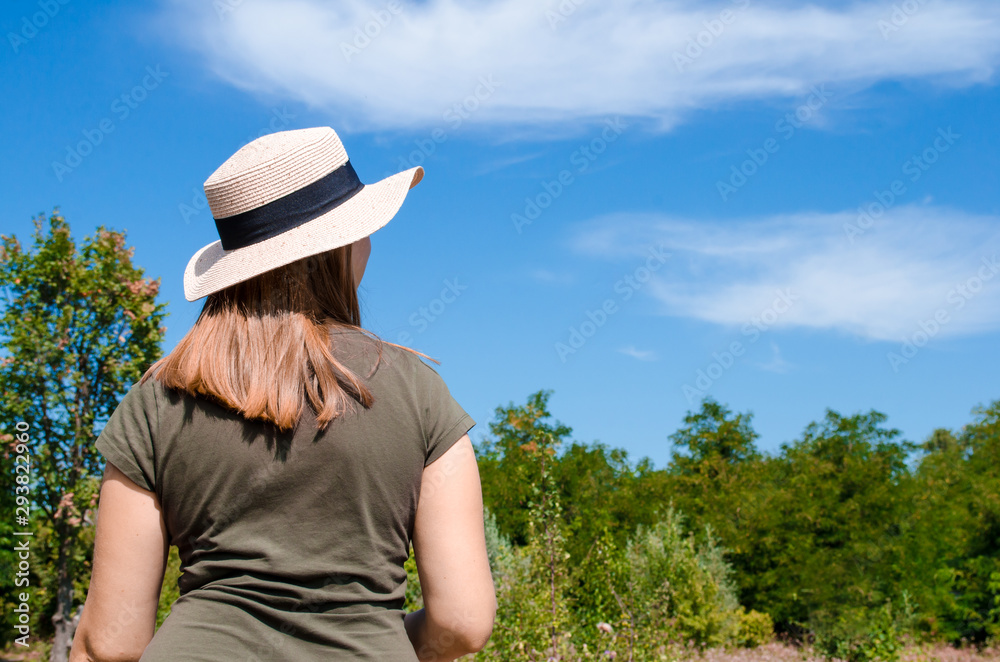 Young girl with blond hair in gray dress and light hat, turned her back, on a background of blue sky, trees and clouds