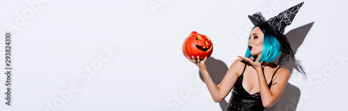 panoramic shot of sexy girl in black witch Halloween costume with blue hair blowing kiss to spooky carved pumpkin on white background photo