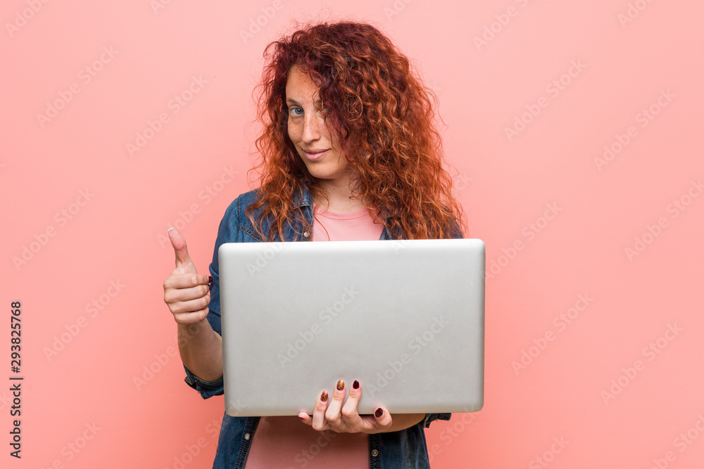 Young caucasian redhead woman holding a laptop smiling and raising thumb up