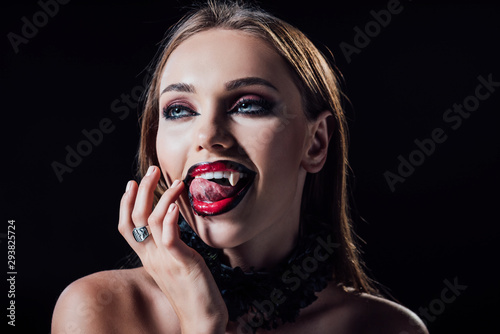 naked scary vampire girl with fangs licking fingers isolated on black photo
