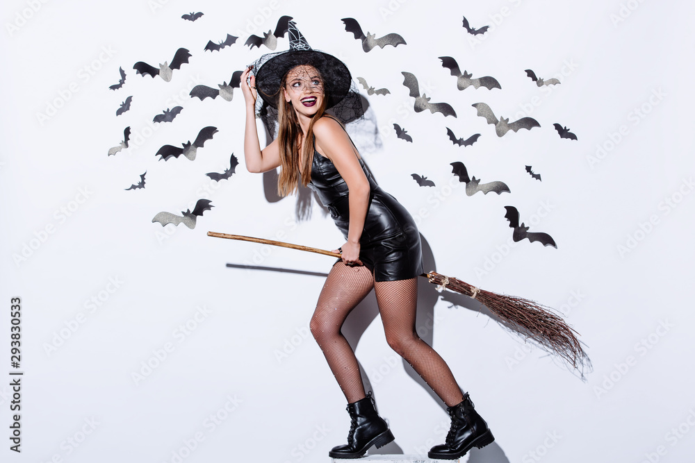 happy girl in black witch Halloween costume on broom near white wall with decorative bats happy girl in black witch Halloween costume on broom near white wall with decorative bats