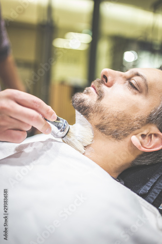 Fashionable man client during beard shaving in barber shop.