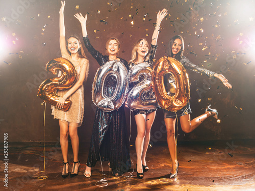 Beautiful Women Celebrating New Year. Happy Gorgeous Girls In Stylish Sexy Party Dresses Holding Gold and Silver 2020 Balloons, Having Fun At New Year's Eve Party. Holiday Celebration.Raising hands