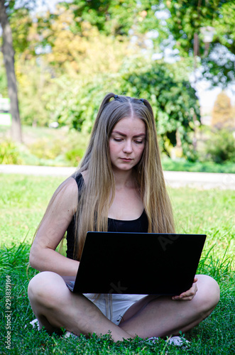 girl with a laptop on the grass sits in summer clothes on a sunny day