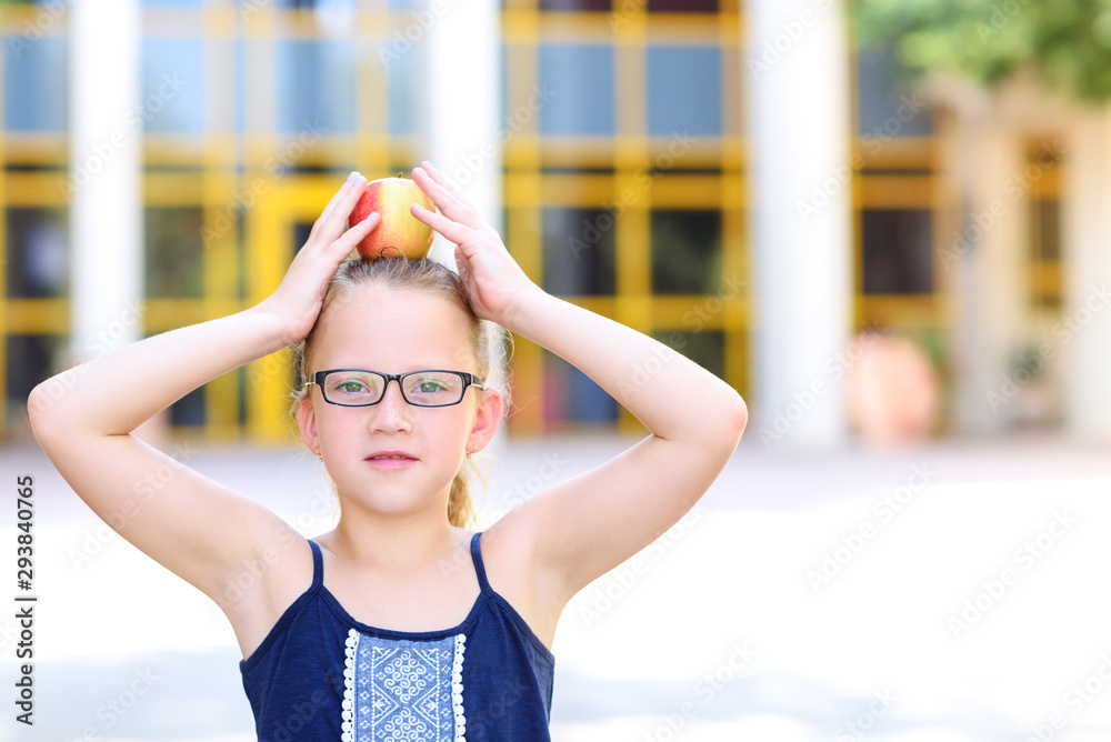 Happy Girl In Eyeglasses With Apple on Her Head. Great Portrait Of School Pupil Outside Classroom. Back to school, balance, success, target and healthy eating for child concept.