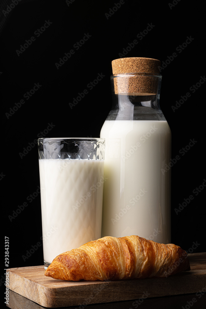 A glass with milk, a bottle with milk and one french croissant on a wooden board. Black background. Glass bottle. Natural cork. White drink. Cow's milk. Classic french croissant. Homemade baking. A glass with milk, a bottle with milk and one french croissant on a wooden board. Black background. Glass bottle. Natural cork. White drink. Cow's milk. Classic french croissant. Homemade baking.