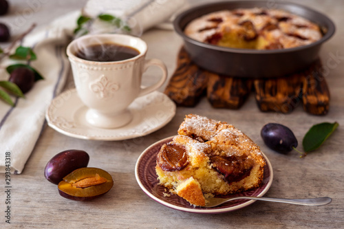 A piece of plum pie on a plate, tea in a cup, plums on a light timbered background.