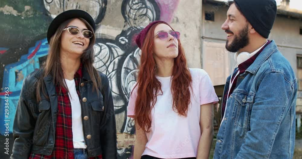 Two Caucasian beautiful and stylish young women and handsome guy, hipsters friends talking and laughing while standing at the graffity on the wall. Outdoor. Two Caucasian beautiful and stylish young women and handsome guy, hipsters friends talking and laughing while standing at the graffity on the wall. Outdoor.