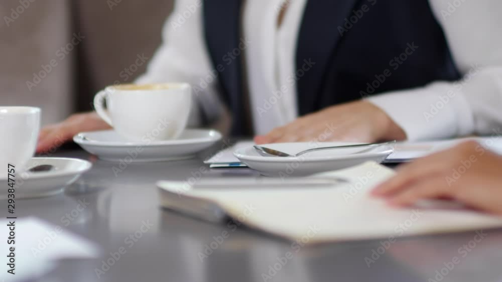 Close-up view of hands of unrecognizable businesswoman writing down in notepad and drinking coffee while sitting with colleagues at cafe table Close-up view of hands of unrecognizable businesswoman writing down in notepad and drinking coffee while sitting with colleagues at cafe table