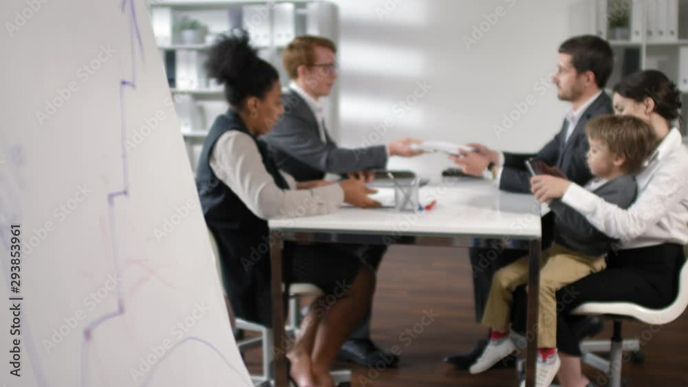 Rack focused shot of multiethnic team of business people discussing project at office table while businesswoman sitting with little son on her knees and playing on digital tablet Rack focused shot of multiethnic team of business people discussing project at office table while businesswoman sitting with little son on her knees and playing on digital tablet