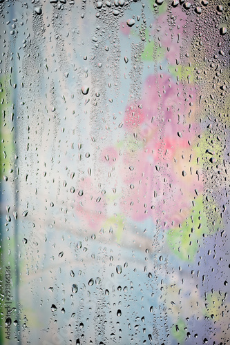 Drops of water flow down the surface of a transparent glass on an abstract blurred bright colored background.